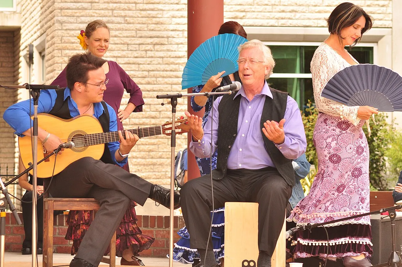 A flamenco group playing outdoors: a seated guitarist, a singer on a cajón and two dancers behind them holding fans