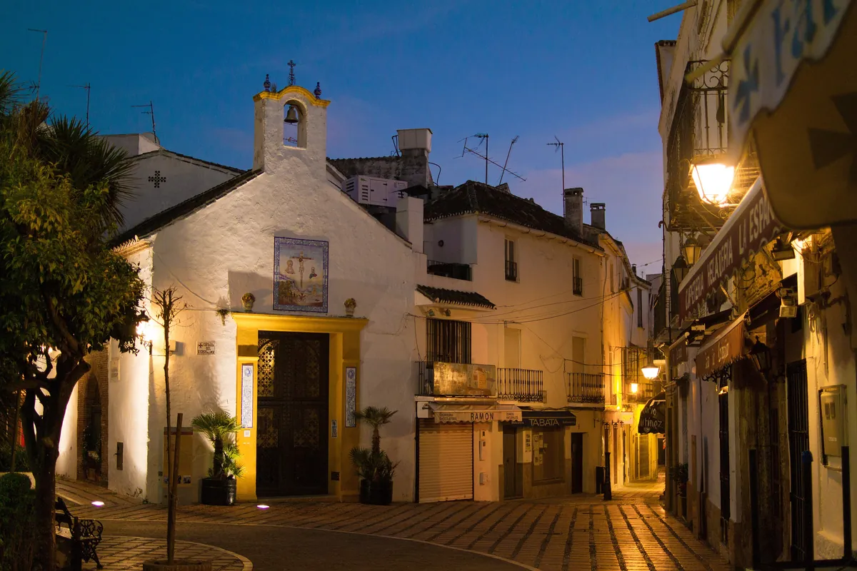 A small whitewashed chapel on a lane in Marbella's old town at dusk, with warm street lamps lit and a deep blue sky above
