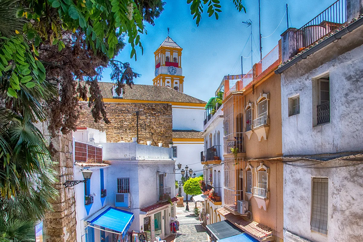 A steep lane in Marbella's old town with colourful houses, a blue awning, a church bell tower with a yellow dome in the distance and a green tree overhead