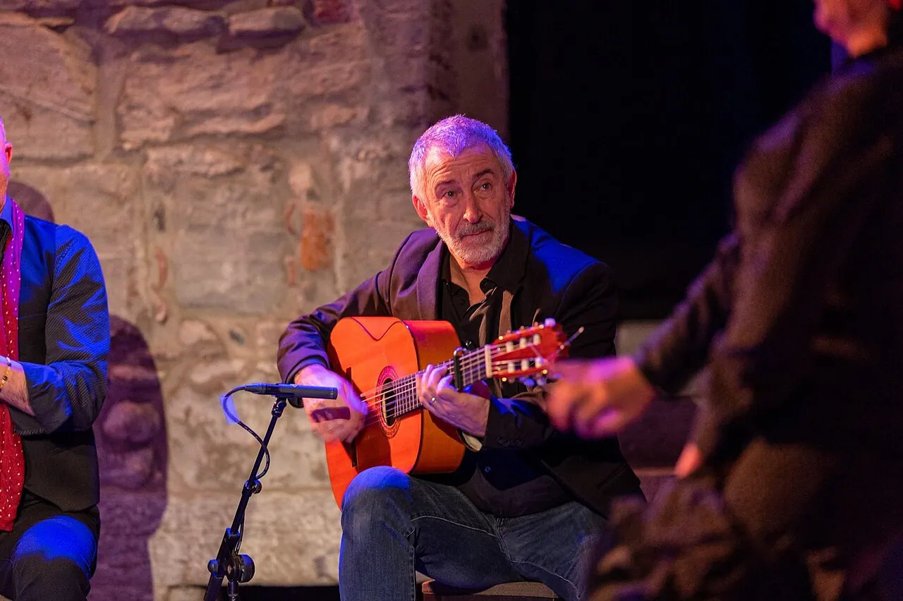 A flamenco guitarist in a dark jacket seated under stage light, playing a guitar with an orange top, with a stone wall behind him