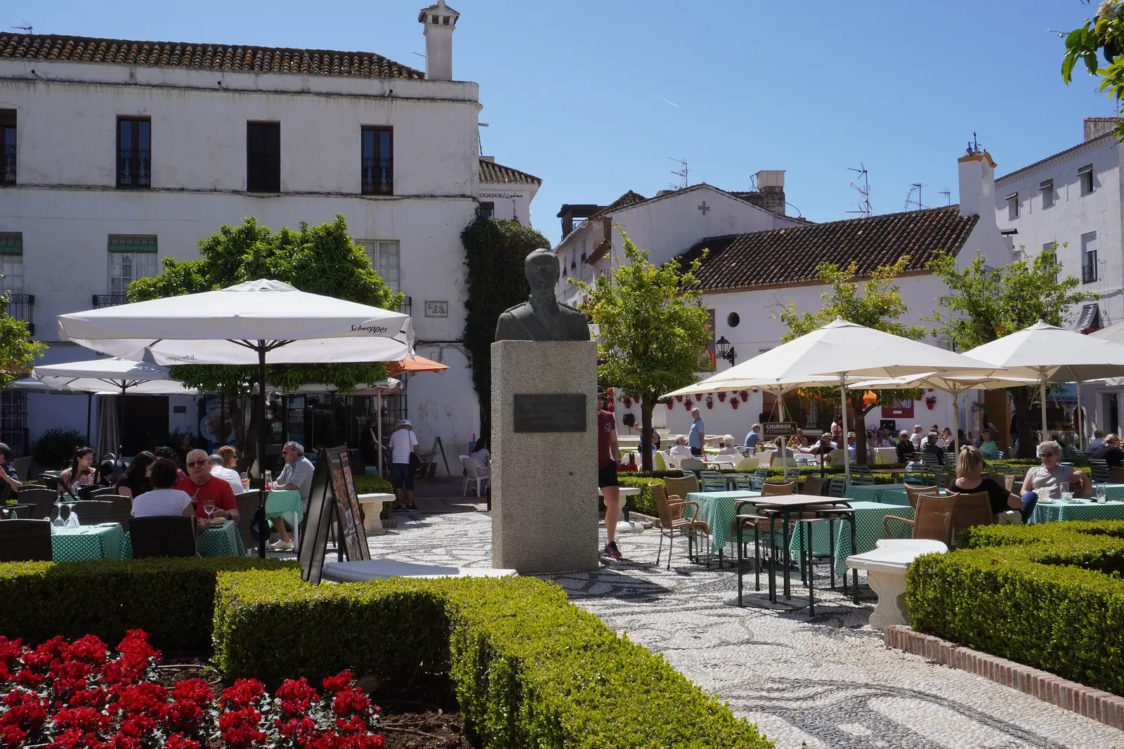 A sunlit square in Marbella's old town with café terraces under white parasols, clipped hedges, red flowers and whitewashed buildings